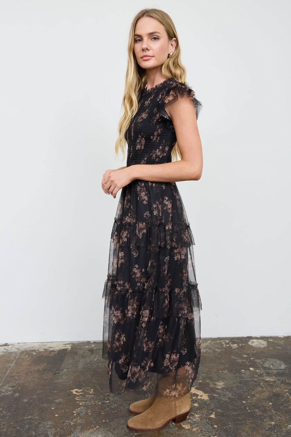 A woman modeling a sleeveless black tulle midi dress with a brown floral pattern, paired with brown suede boots, standing against a white wall.