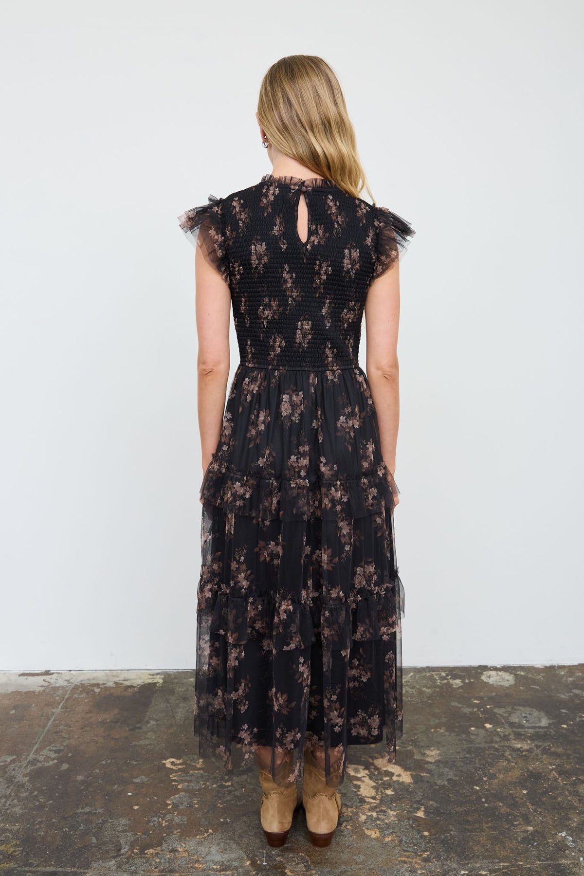 A woman viewed from behind wearing a black midi dress with a brown floral pattern, sheer tulle cap sleeves, and a keyhole back detail, standing on a rustic concrete floor against a white wall.