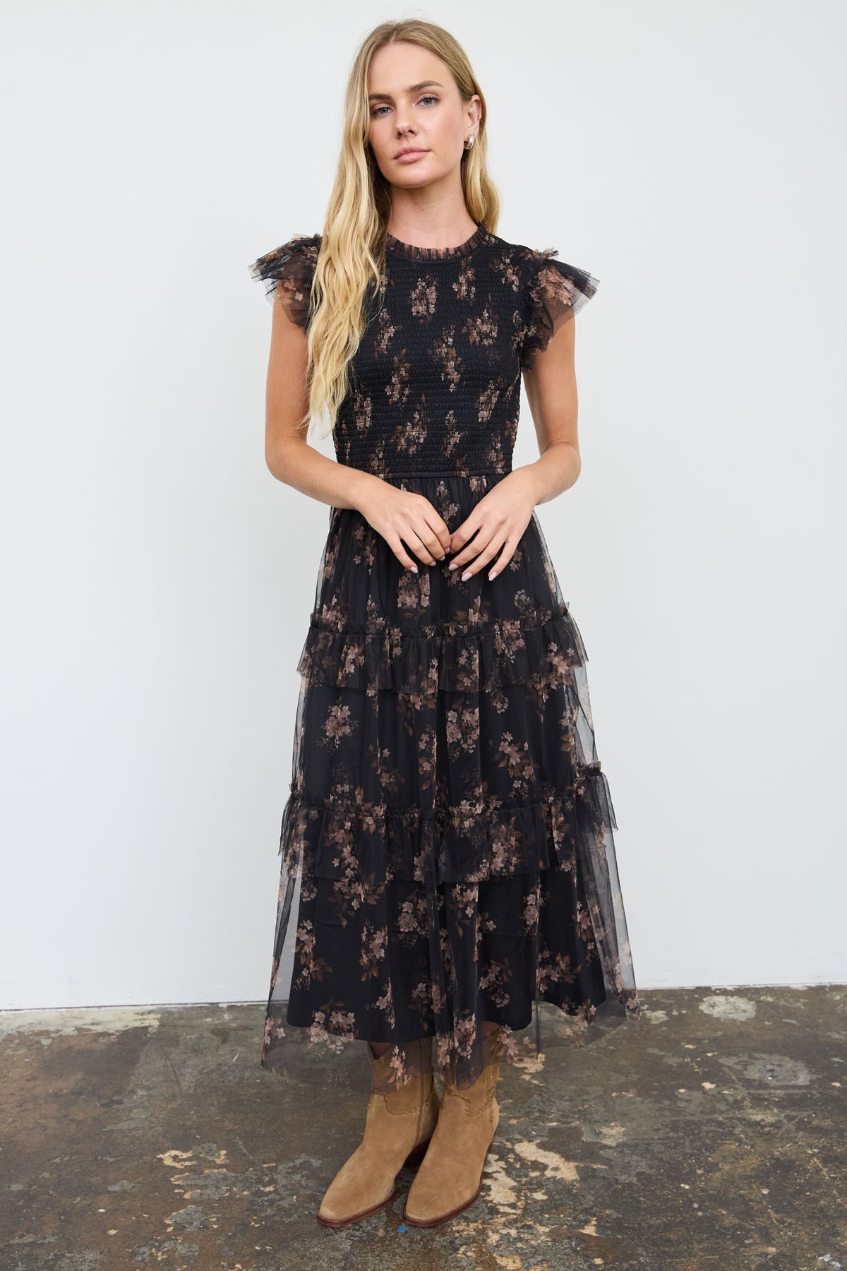 A woman modeling a black tulle midi dress with a brown floral pattern, ruffled cap sleeves, and brown suede ankle boots, standing against a white wall.