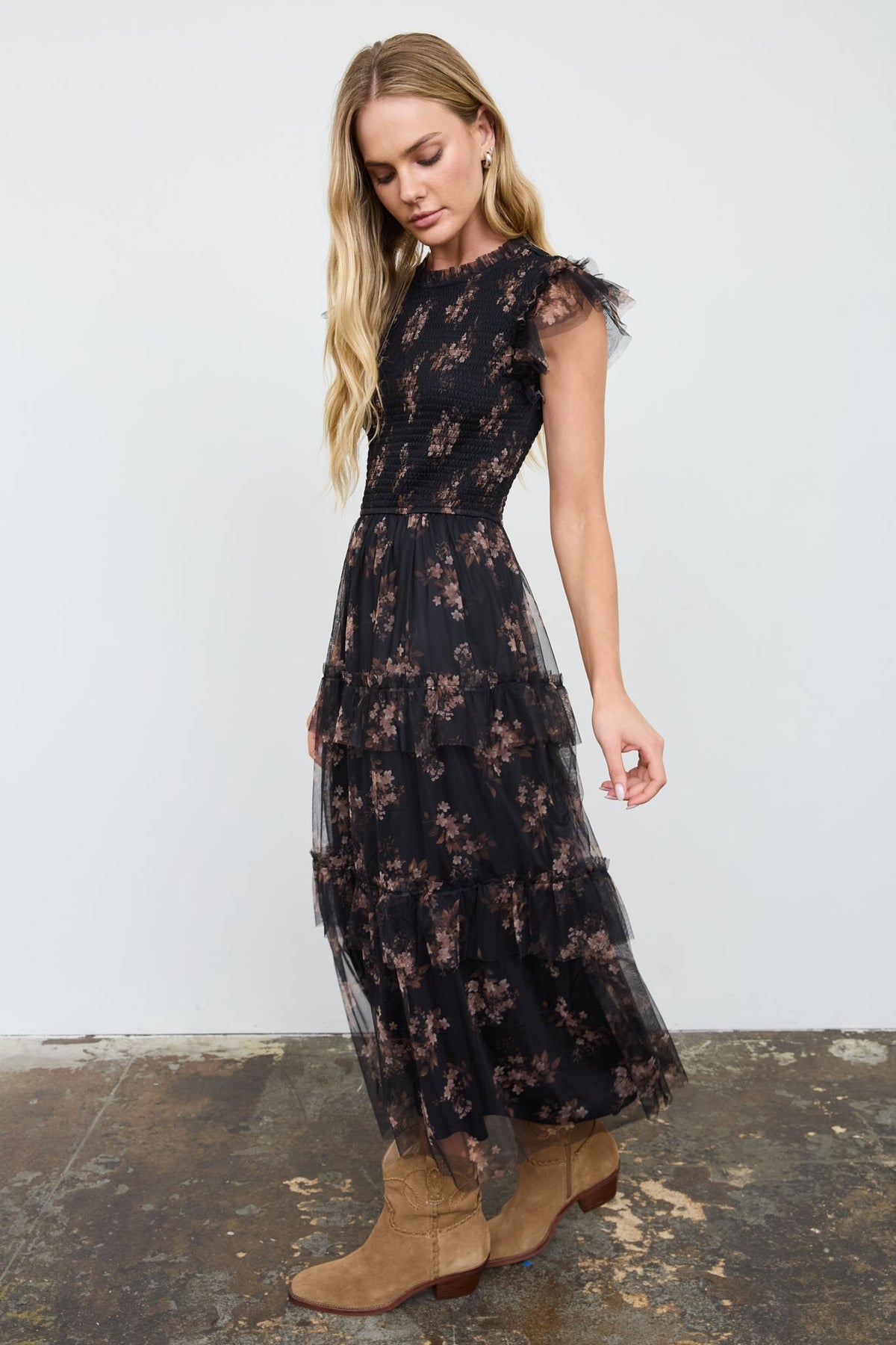 A woman modeling a black tiered tulle maxi dress with a brown floral pattern and sheer flutter sleeves, paired with tan suede ankle boots, set against a plain white wall and concrete floor.