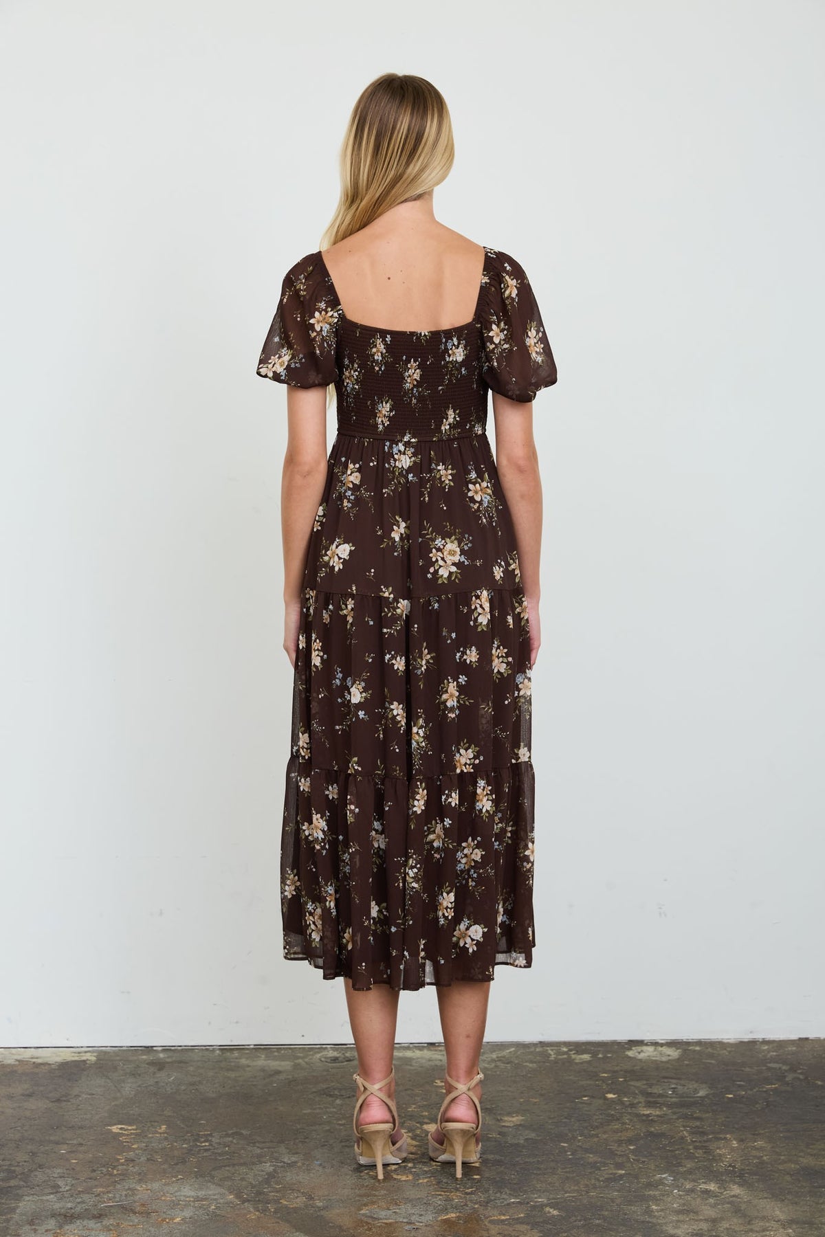 A woman stands with her back to the camera wearing a dark brown midi dress with a beige and white floral pattern, short puff sleeves, and a square neckline. She is wearing beige high-heeled sandals against a plain white wall and concrete floor.