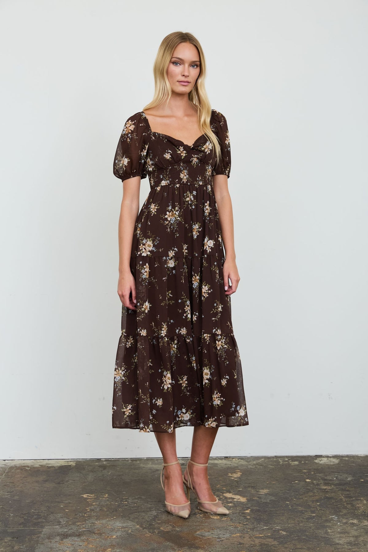 A woman modeling a dark brown midi dress with a beige and white floral print, featuring puff sleeves and a sweetheart neckline, standing against a plain white wall.