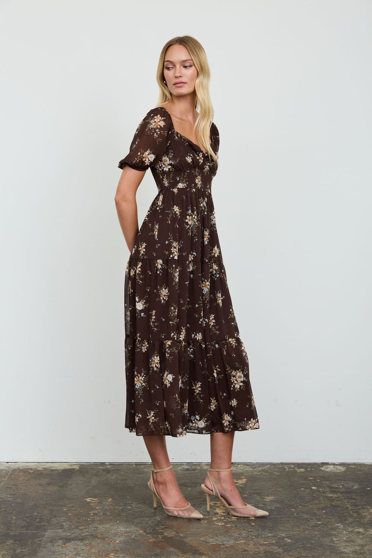 A woman modeling a dark brown midi dress with a beige and white floral pattern, standing against a plain light gray wall.