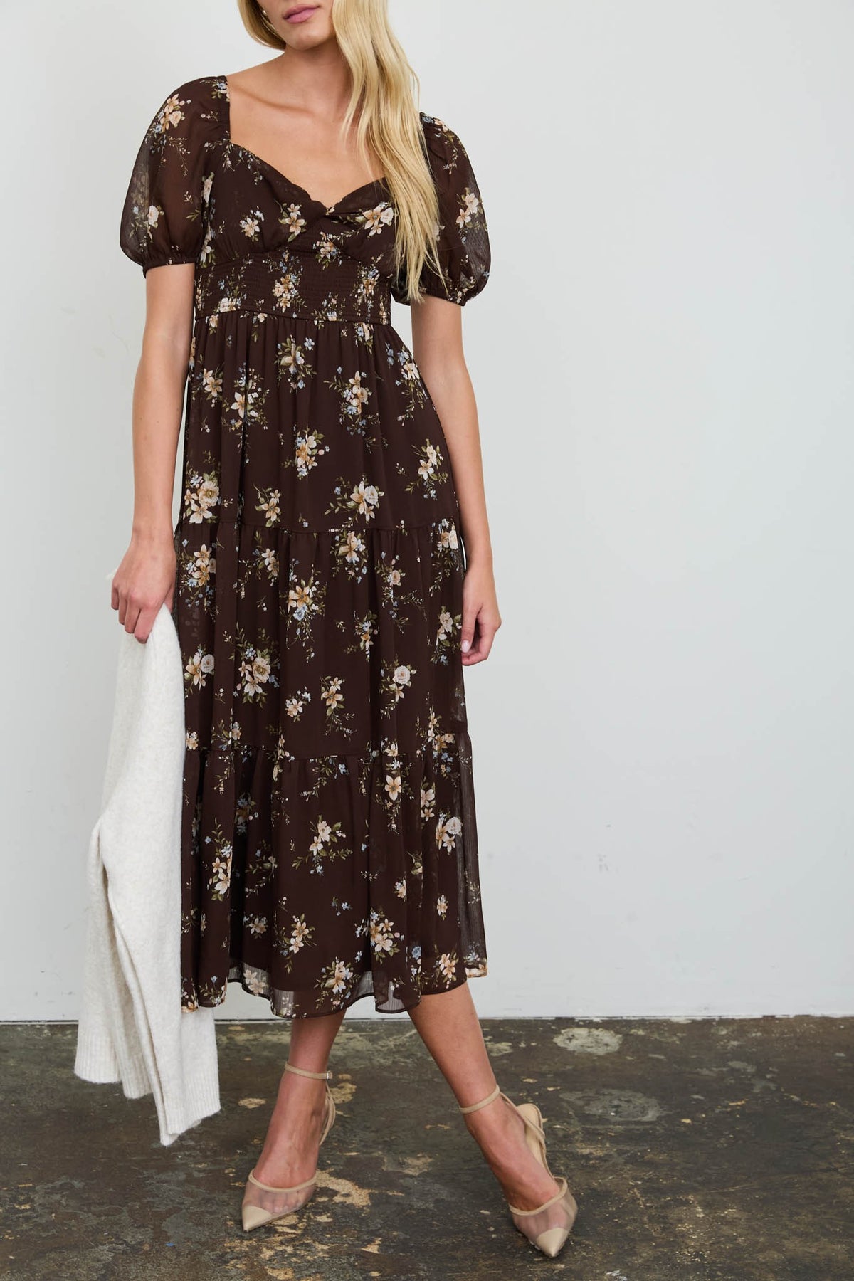 A woman models a dark brown midi dress with a floral print, featuring short puff sleeves and a sweetheart neckline. She is holding a white draped fabric accessory and wearing beige pointed-toe heels against a plain white wall.