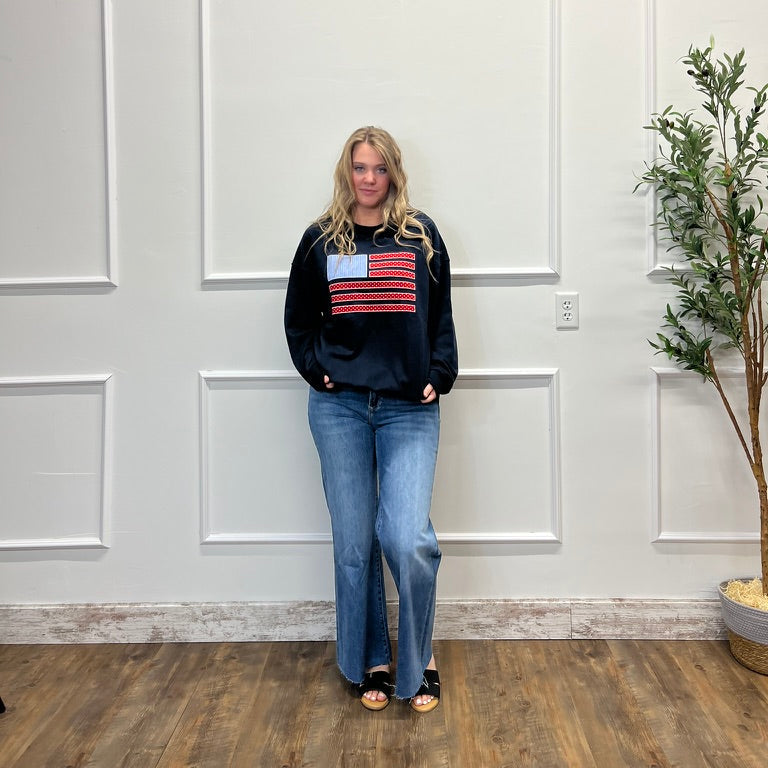 Person wearing a black sweater with an American flag design in a room with white walls and a plant.