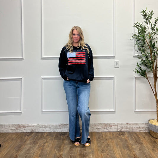 Person wearing a black sweater with an American flag design in a room with white walls and a plant.