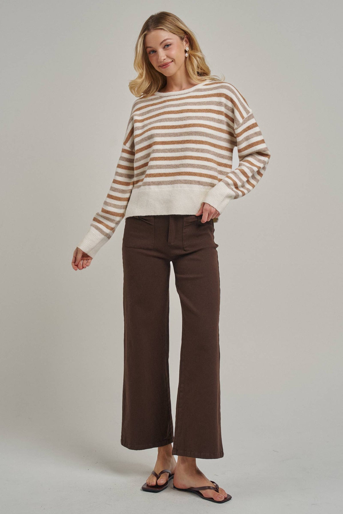 A woman modeling a long-sleeve knit sweater with horizontal tan and cream stripes, paired with dark brown wide-leg cropped pants and black flip-flop sandals, against a light gray studio background.