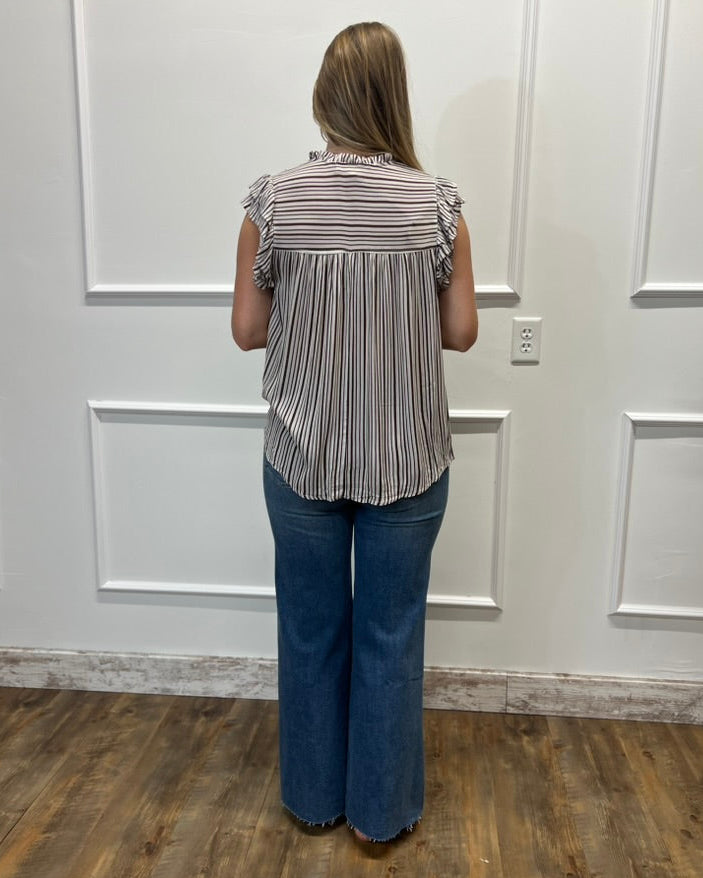 A woman viewed from behind wearing a sleeveless striped blouse with ruffle details and blue wide-leg jeans, standing in front of a white paneled wall on a wood floor.
