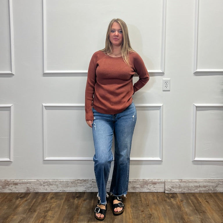 A woman stands indoors against a white paneled wall wearing a rust-brown long-sleeve knit top, blue jeans, and black sandals.