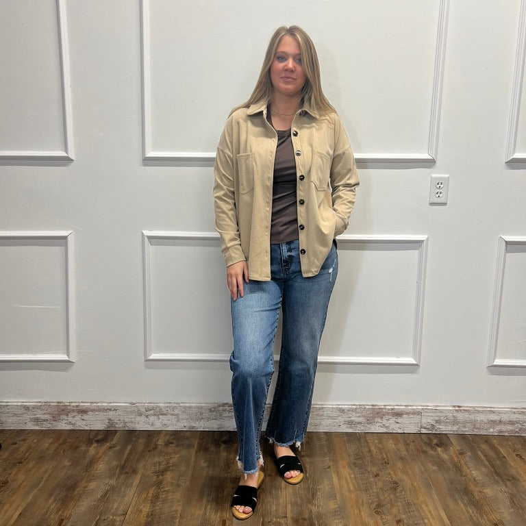 A woman stands in a room with white paneled walls and a wood floor, wearing a beige button-up overshirt over a dark striped top, blue jeans, and black slide sandals.