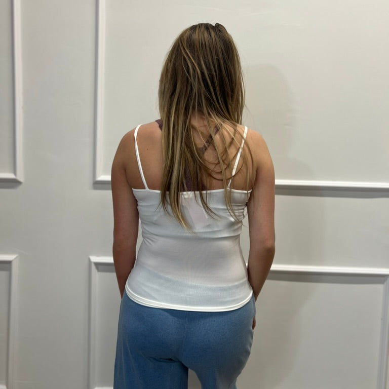 A woman viewed from behind wearing a white spaghetti-strap camisole tank top and blue wide-leg sweatpants, standing against a white paneled wall.