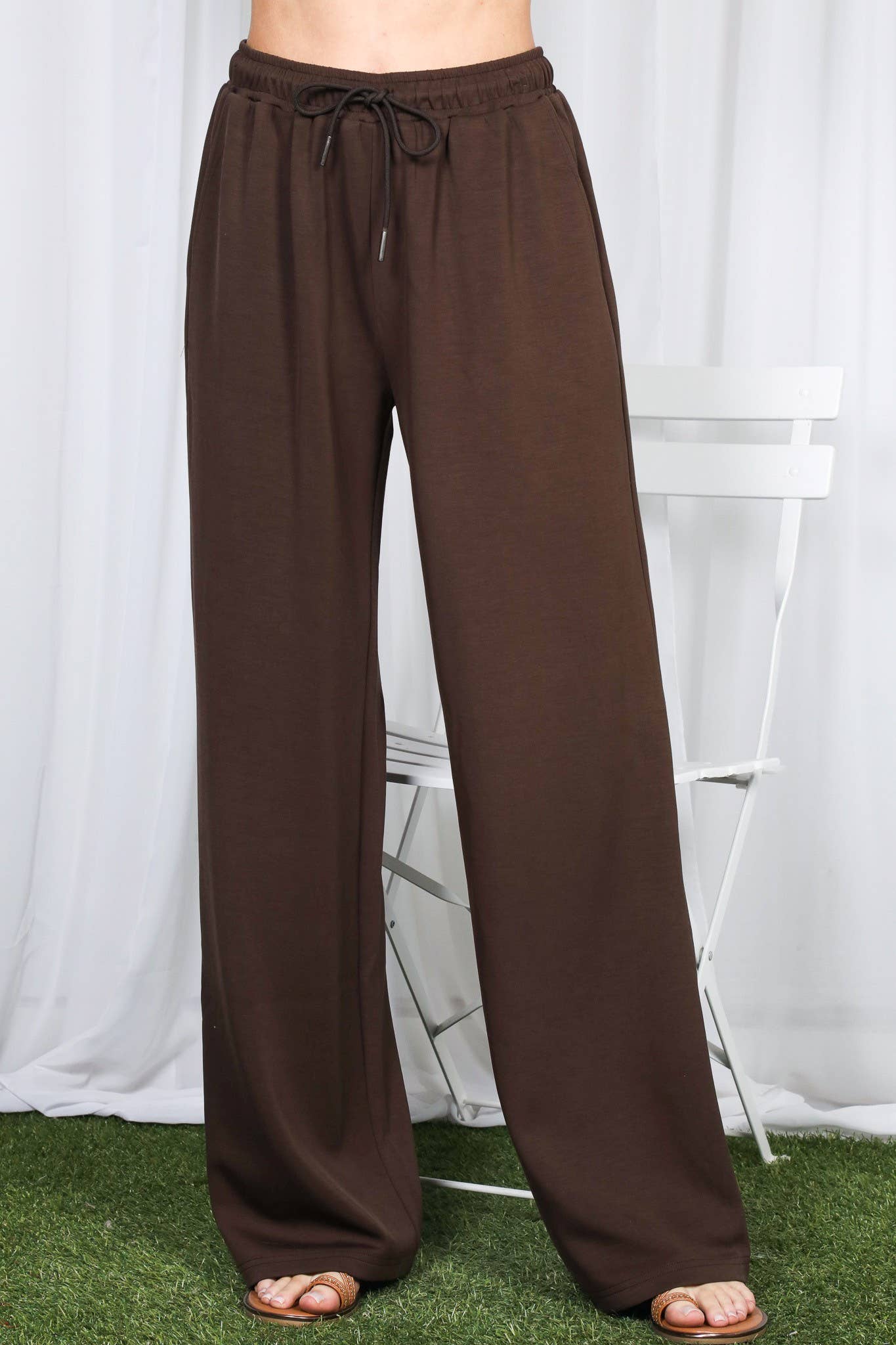 A person wearing loose, wide-leg brown drawstring pants with sandals, standing in front of a white curtain backdrop with a white chair and artificial grass visible.