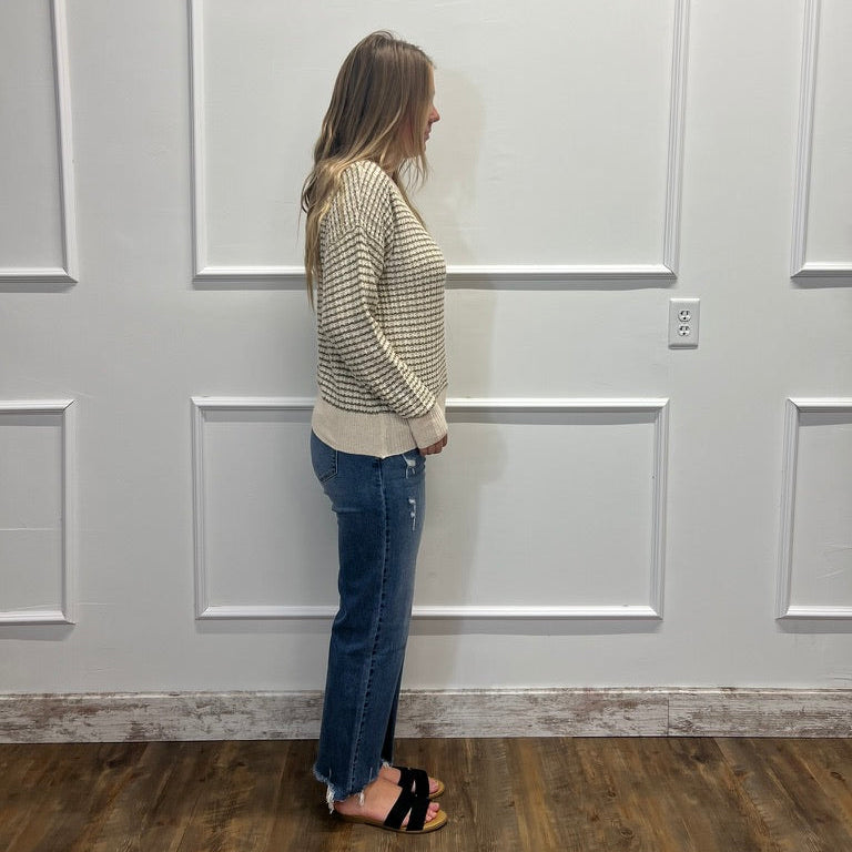 A woman stands in profile wearing a cream and black patterned knit sweater, blue jeans, and black slide sandals, standing against a white paneled wall on a wood floor.