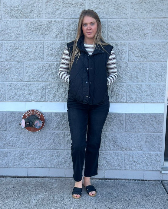 A woman stands outdoors against a light gray stone wall, wearing a black quilted vest over a black and white striped long-sleeve shirt, black pants, and black slide sandals.