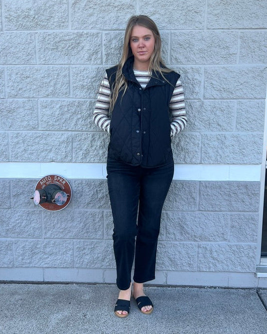 A woman stands outdoors against a light gray stone wall, wearing a black quilted vest over a black and white striped long-sleeve shirt, black pants, and black slide sandals.