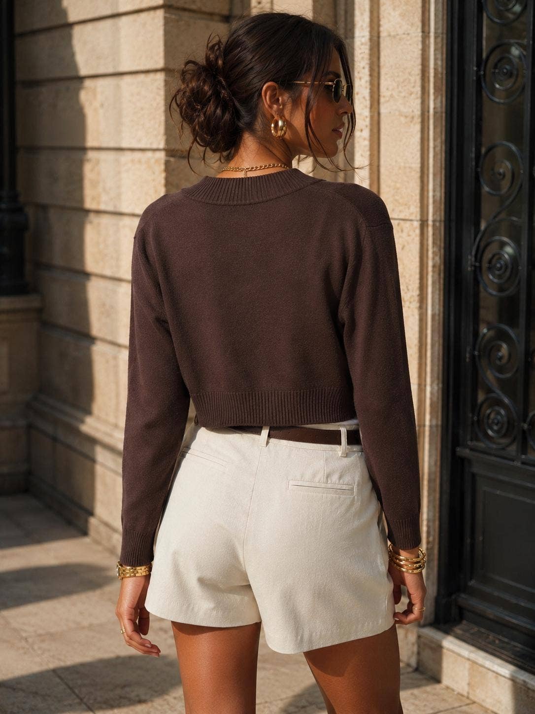 A woman wearing a dark brown long-sleeve cropped knit sweater and off-white shorts, photographed from behind in an outdoor urban setting with a stone building and black metal door in the background.