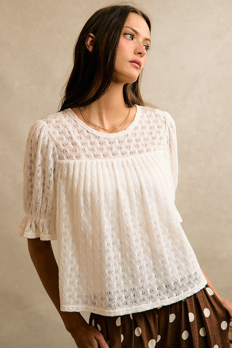 A woman wearing a white eyelet lace short-sleeve blouse with a gathered bust, paired with a brown and white polka dot skirt, posed against a neutral beige textured wall.