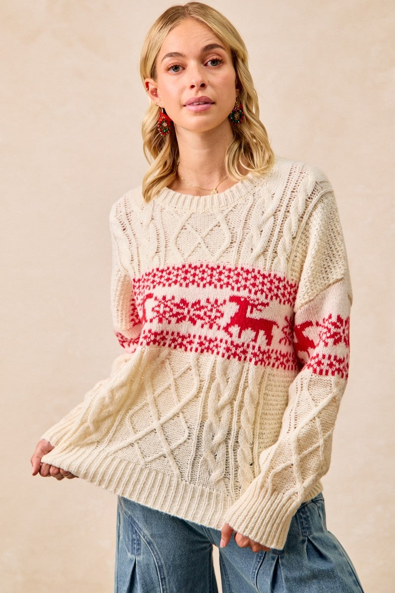 A woman wearing a cream cable-knit sweater with a red Nordic-style holiday pattern featuring reindeer and snowflakes, paired with blue jeans against a neutral beige background.