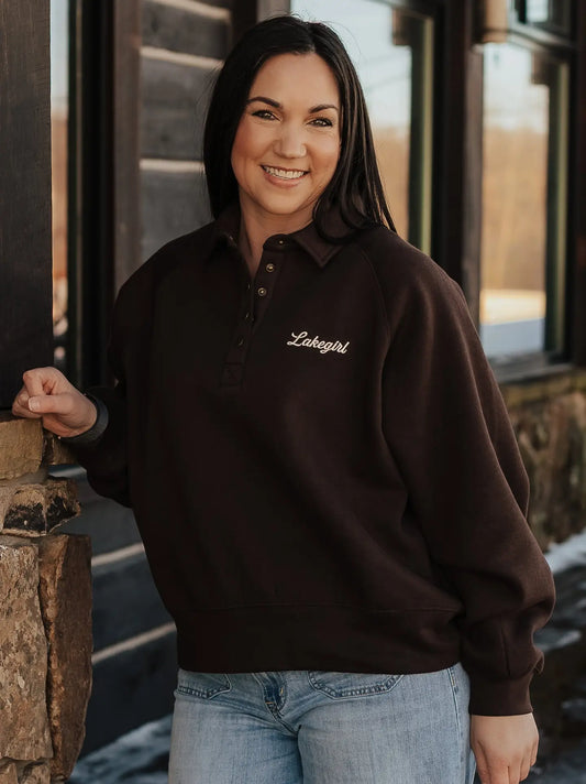 A woman wearing a dark brown long-sleeve polo sweatshirt with 'Lakegirl' embroidered in white script on the left chest, paired with light blue jeans, standing outdoors in front of rustic wooden buildings.