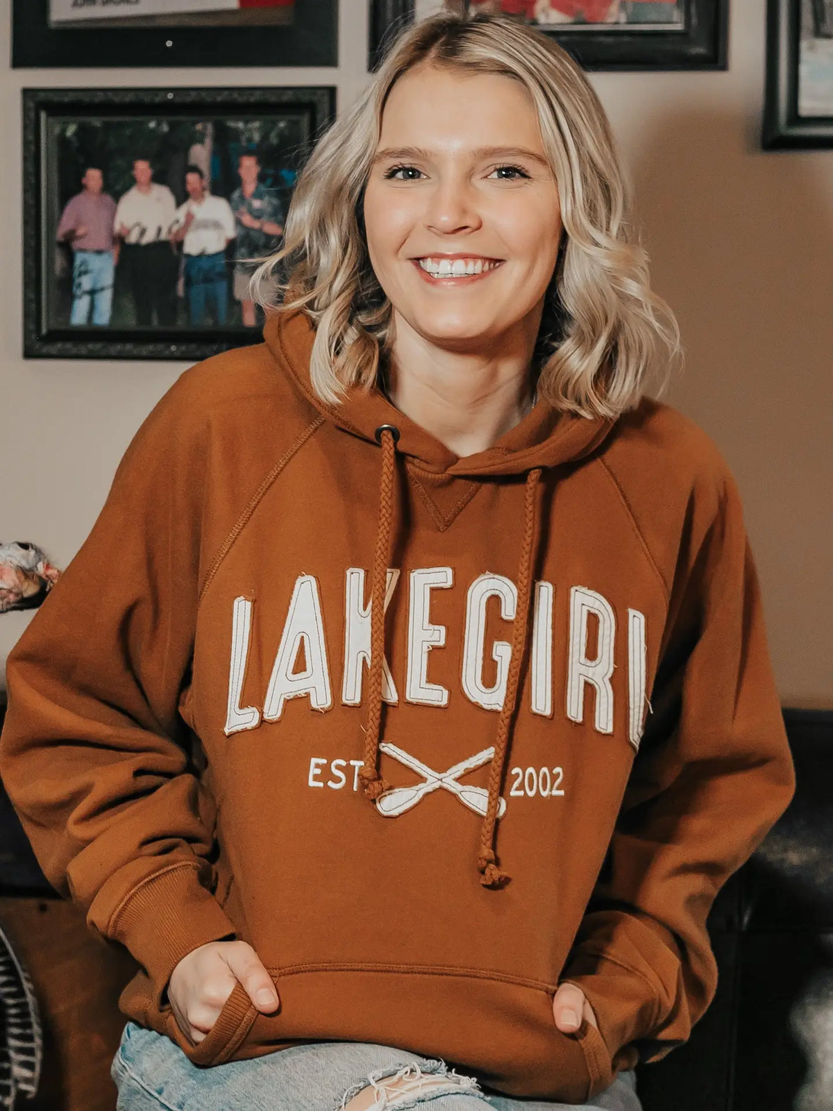A smiling blonde woman wearing a rust-brown pullover hoodie with 'LAKEGIRL EST. 2002' printed in white across the front, seated indoors with framed photos on the wall behind her.