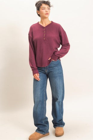 A model wearing a long-sleeve henley shirt in a deep burgundy/maroon color, paired with blue jeans and tan suede boots, standing against a plain off-white studio background.