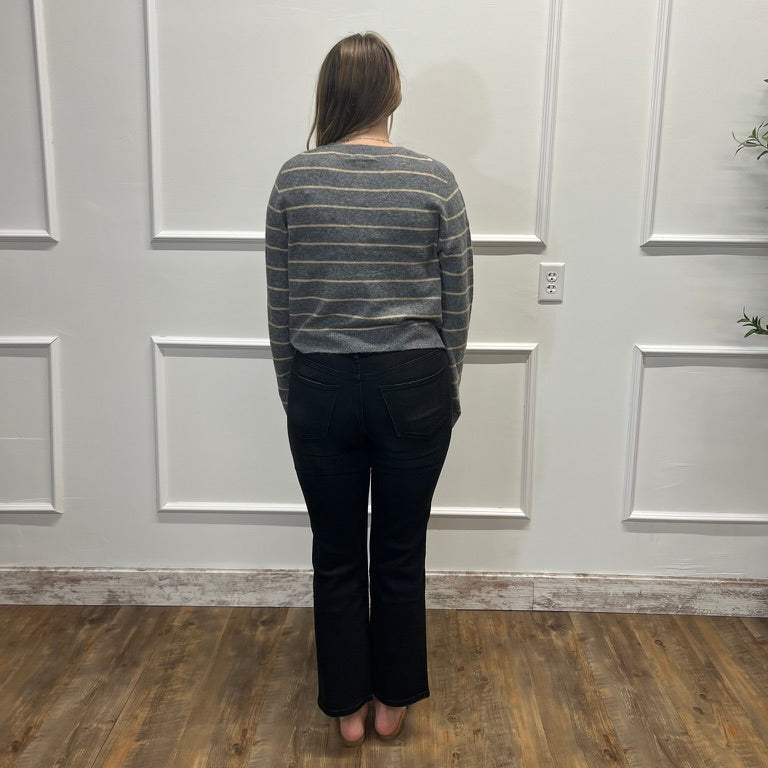 A woman stands with her back to the camera wearing a gray and white striped long-sleeve knit sweater and black pants, standing on a wood floor against a white paneled wall.