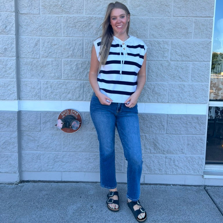 A woman stands against a light gray stone wall wearing a sleeveless top with bold black and white horizontal stripes, blue jeans, and black sandals.