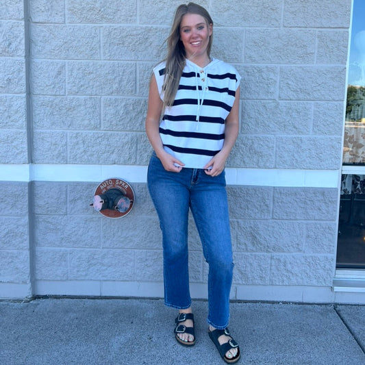 A woman stands against a light gray stone wall wearing a sleeveless top with bold black and white horizontal stripes, blue jeans, and black sandals.