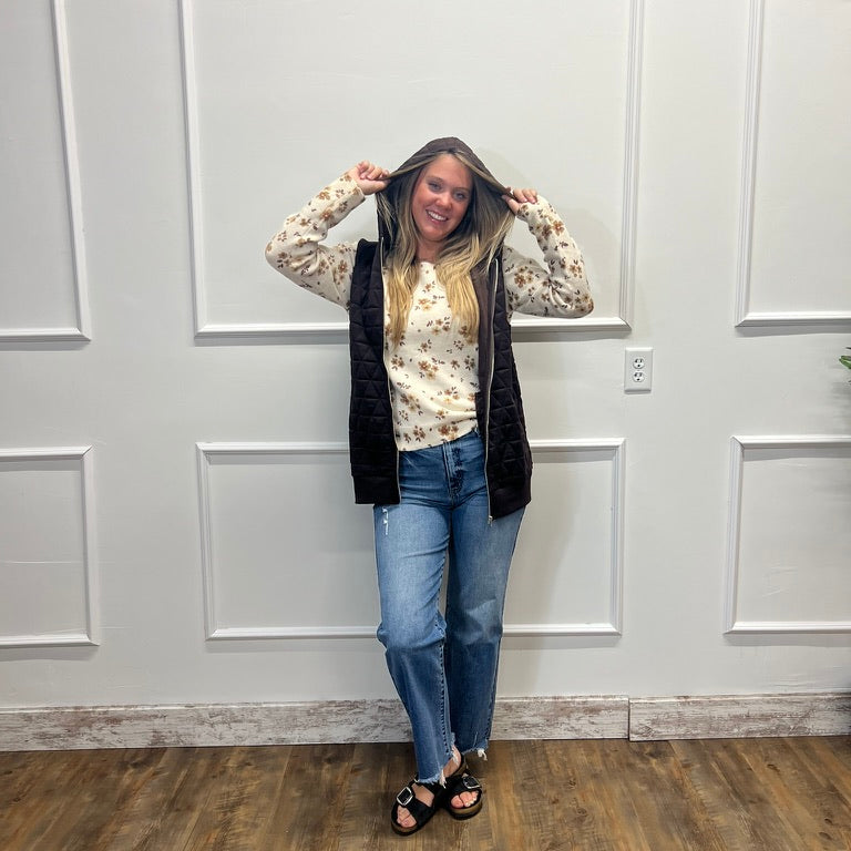 A woman stands in a room with white paneled walls and a wood floor, wearing a cream floral long-sleeve shirt, a black sleeveless vest, blue jeans, and black sandals.