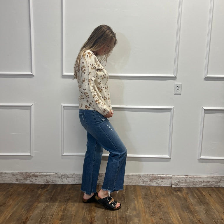 A woman stands in profile wearing a cream floral long-sleeve top, blue cropped flare jeans, and black slide sandals, standing against a white paneled wall on a wood floor.
