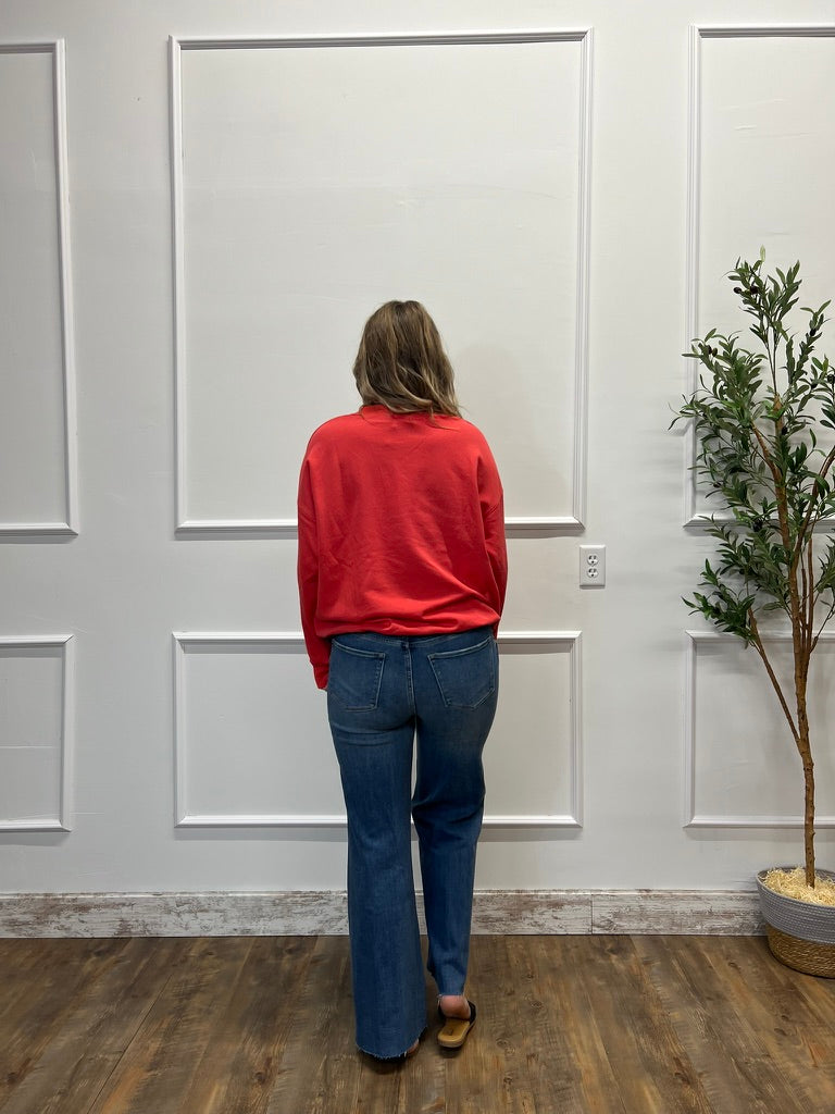Person wearing a red sweater and blue jeans standing in front of a white paneled wall.