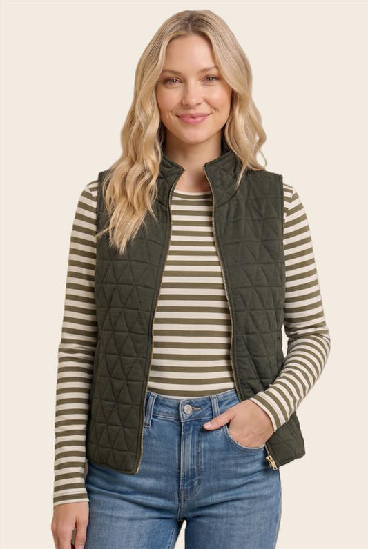 A woman wearing an olive green quilted zip-front vest over a beige and olive striped long-sleeve top, paired with blue jeans, against a plain light beige studio background.