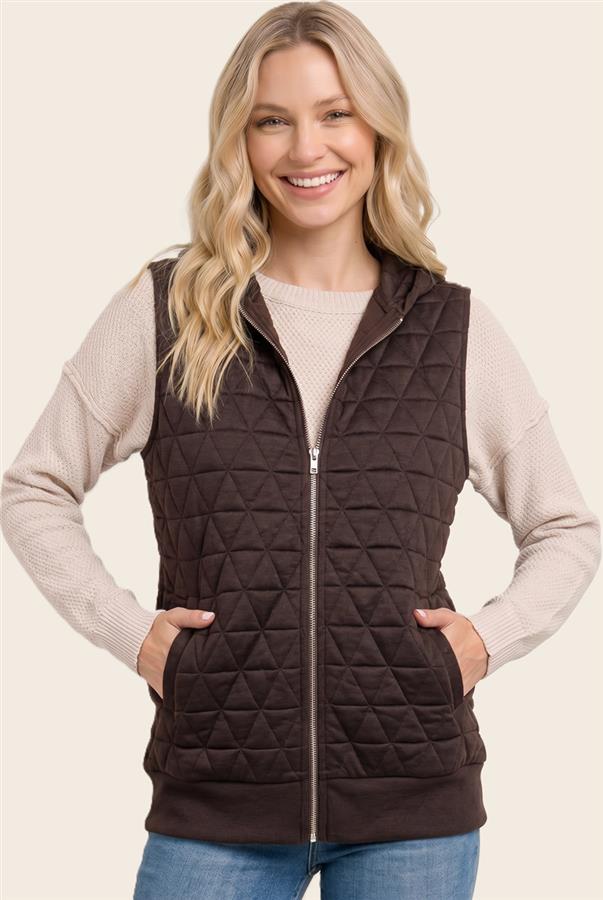 A smiling blonde woman wearing a dark brown diamond-quilted zip-up vest over a beige knit sweater and blue jeans, set against a solid light beige background.