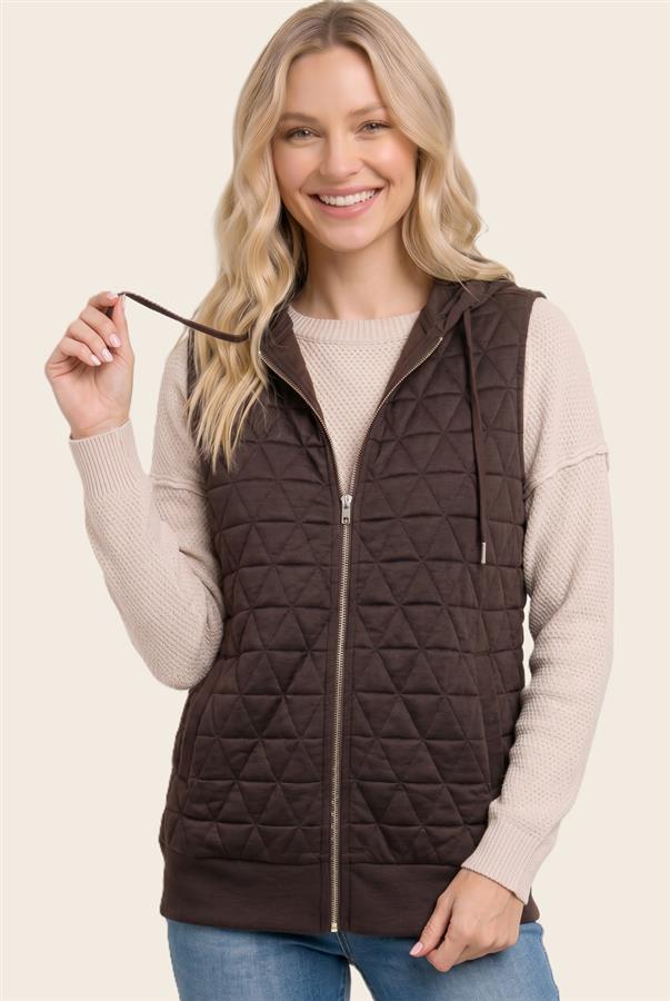 A smiling blonde woman wearing a dark brown diamond-quilted zip-up hooded vest over a beige knit sweater and blue jeans, holding the drawstring against a light beige studio background.