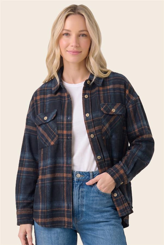 A woman modeling a dark navy and brown plaid button-up shirt jacket over a white t-shirt and blue jeans, set against a solid light beige background.