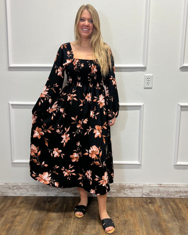A woman stands in a room with white paneled walls and a wood floor, wearing a long-sleeve black maxi dress with a peach and orange floral pattern. She is wearing black slide sandals.