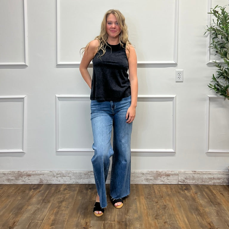 Woman wearing a black sleeveless top and blue jeans standing against a white paneled wall.
