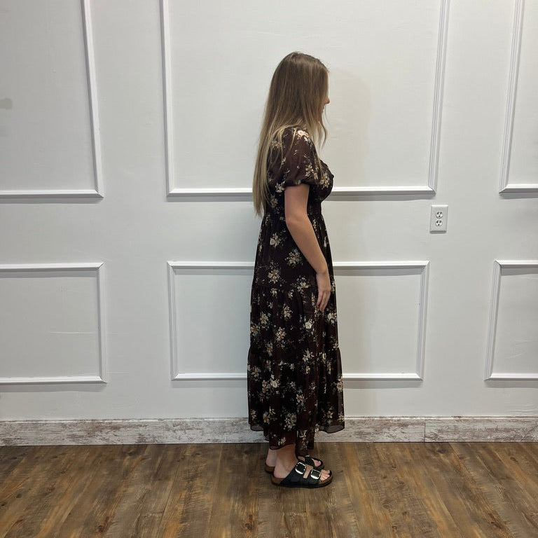 A woman stands in profile wearing a dark brown short-sleeve maxi dress with a beige floral pattern, paired with black slide sandals, set against a white paneled wall and wood-look flooring.
