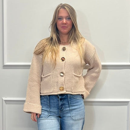 A woman stands in a room with white paneled walls and a wood floor, wearing a beige ribbed cardigan with brown buttons, light blue flare jeans, and brown sandals.