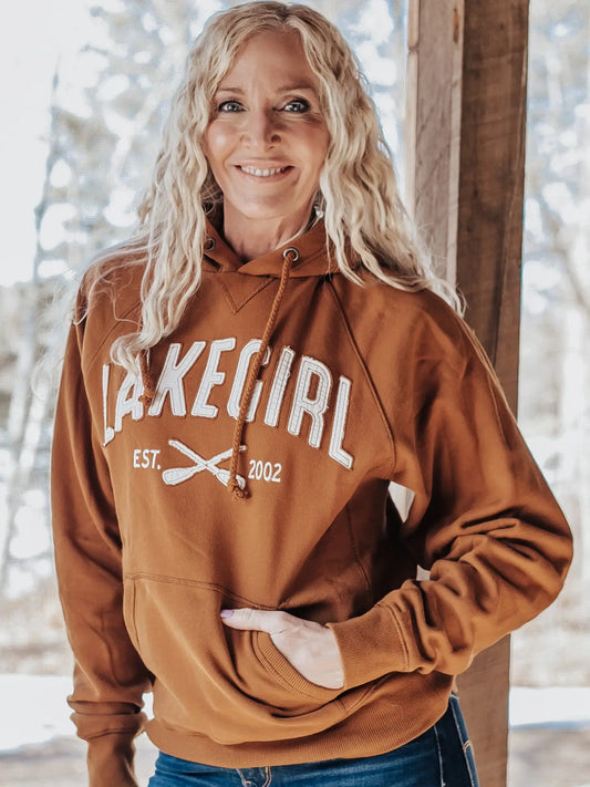 A smiling blonde woman wearing a rust-brown pullover hoodie with 'LAKEGIRL EST. 2002' printed in white across the chest, standing outdoors against a blurred background with trees and a wooden post.