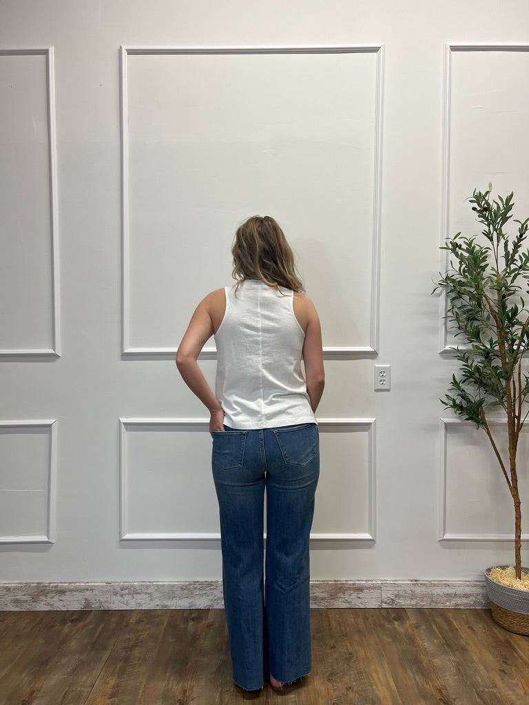 Person wearing a white sleeveless top and blue jeans standing in front of a white paneled wall.