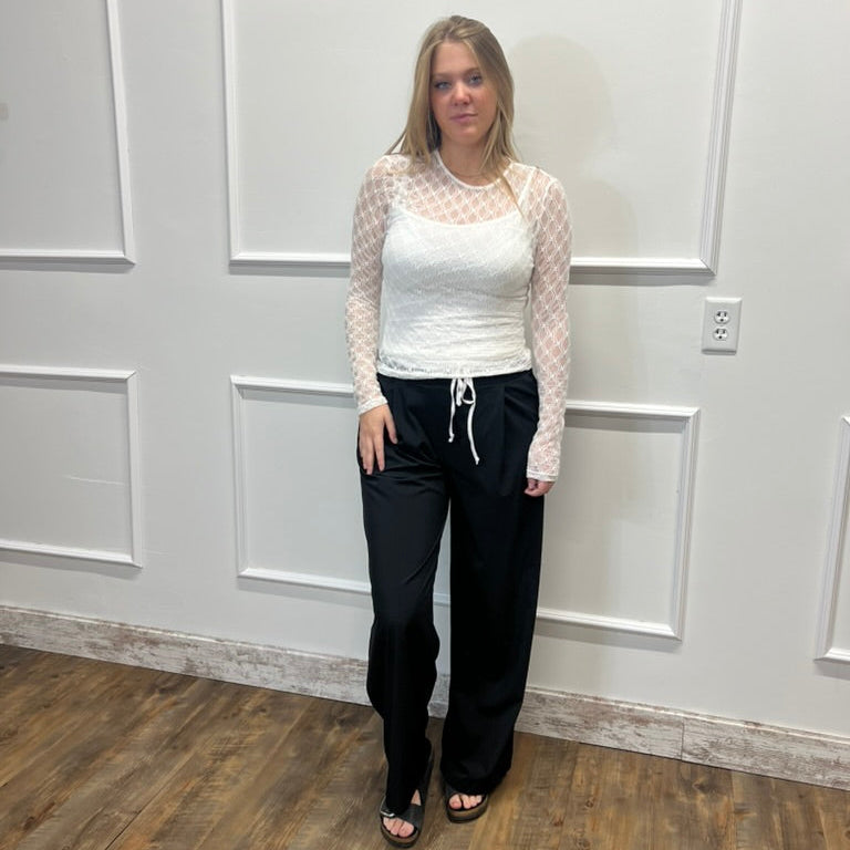 A woman modeling a white long-sleeve textured top with sheer patterned sleeves and black wide-leg drawstring pants, standing in a room with white paneled walls and a wood floor.