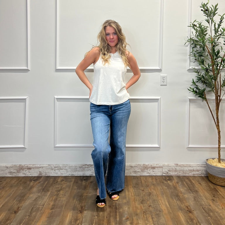 Woman wearing a white sleeveless top and blue jeans standing in a room with a white paneled wall and a plant.