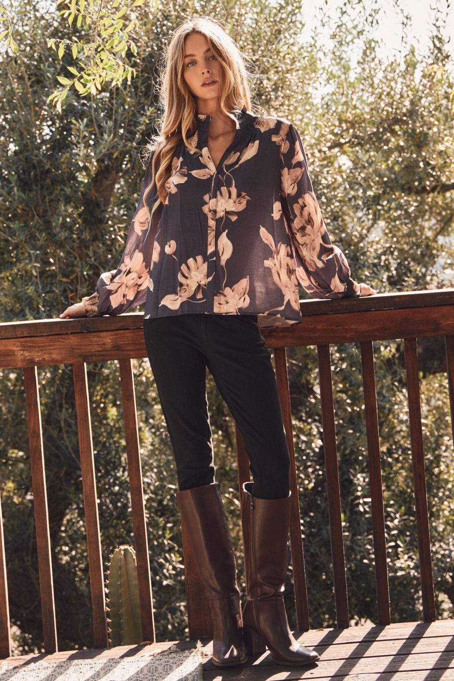 Woman standing on a wooden deck wearing a floral blouse and dark pants, with trees in the background.
