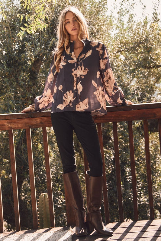 Woman standing on a wooden deck wearing a floral blouse and dark pants, with trees in the background.