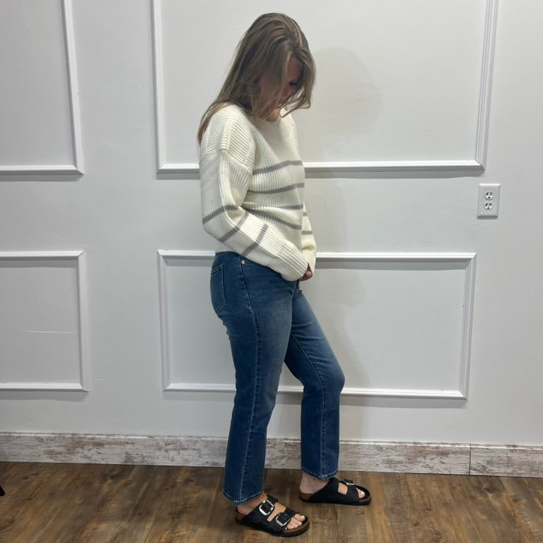 A woman stands in a room with white paneled walls and a wood floor, wearing a cream ribbed knit sweater with horizontal gray stripes, blue jeans, and black slide sandals.