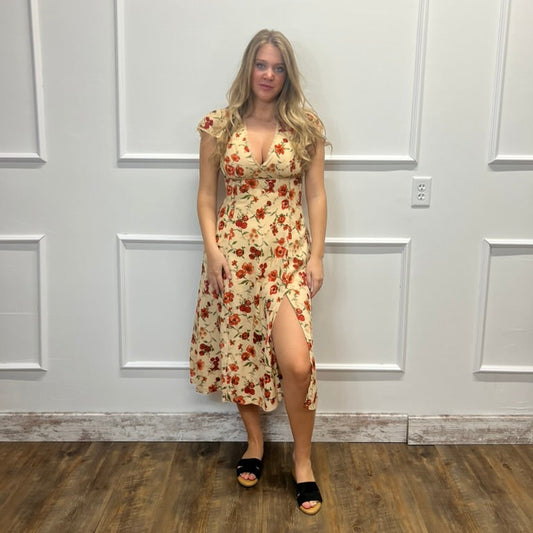 A woman stands in a room with white paneled walls and a wood floor, wearing a sleeveless midi dress with a red and orange floral pattern on a cream background, paired with black slide sandals.