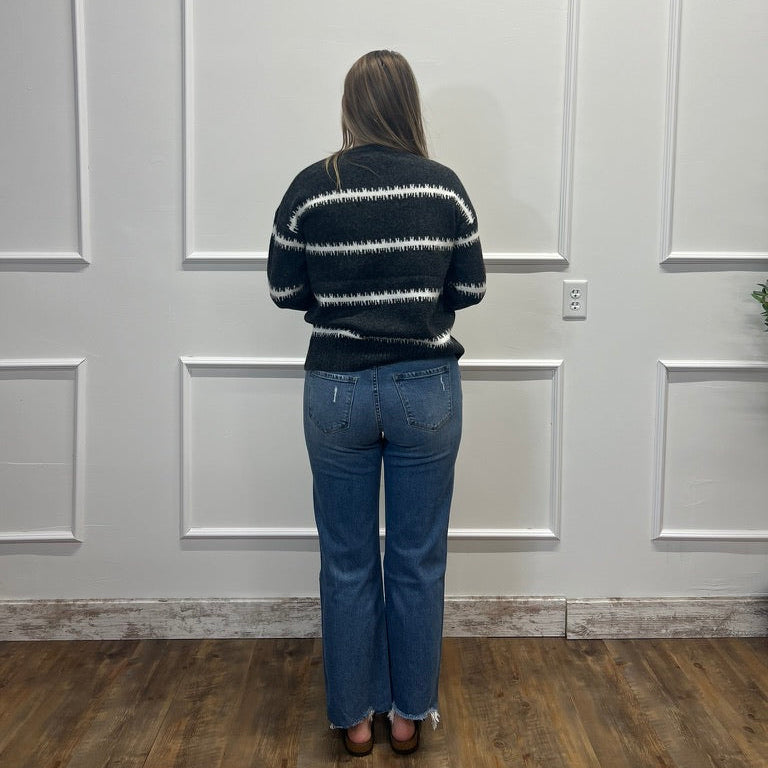 A woman viewed from behind wearing a black knit sweater with white horizontal stripes and blue jeans, standing in front of a white paneled wall on a wood floor.