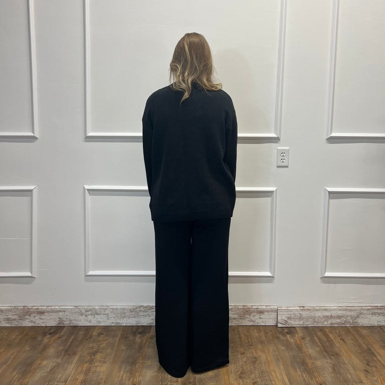 A person standing with their back to the camera, wearing a black long-sleeve top and black wide-leg pants in a room with white paneled walls and a wood floor.