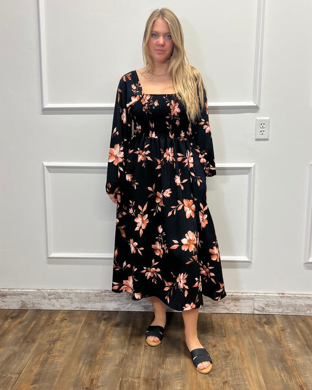 A woman stands in a room with white paneled walls and wood floors, wearing a long black dress with a peach and orange floral pattern and black slide sandals.